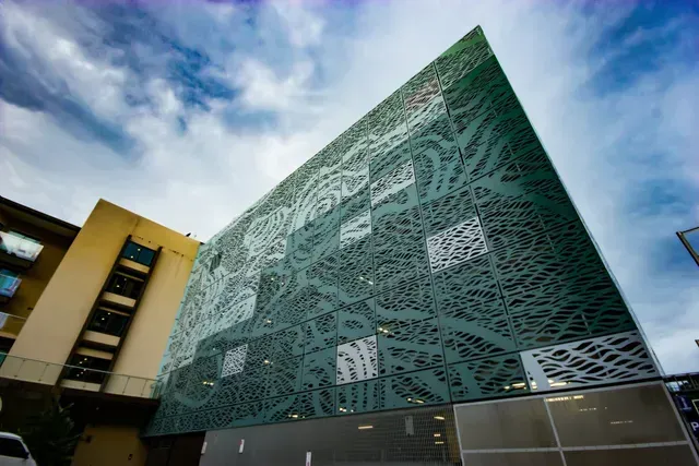 Green building facade with intricate, nature-inspired patterns under a cloudy sky.