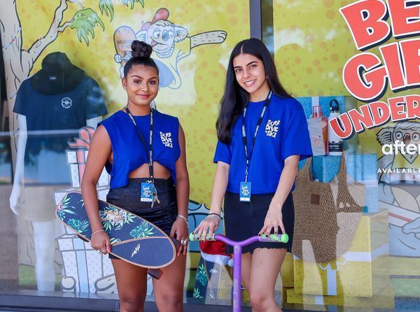 Two women pose in front of a store window; one holds a skateboard, the other a scooter.