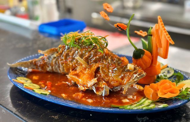 Deep-fried fish with red sauce, decorated with carved carrots, cucumber, and green onion on a blue plate.