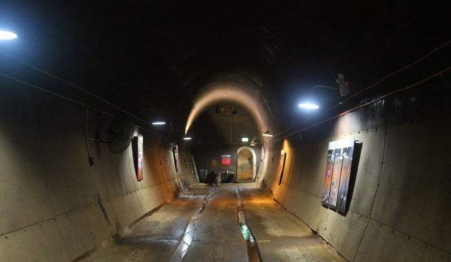 Dark, curved tunnel with overhead lights. Metallic walls.