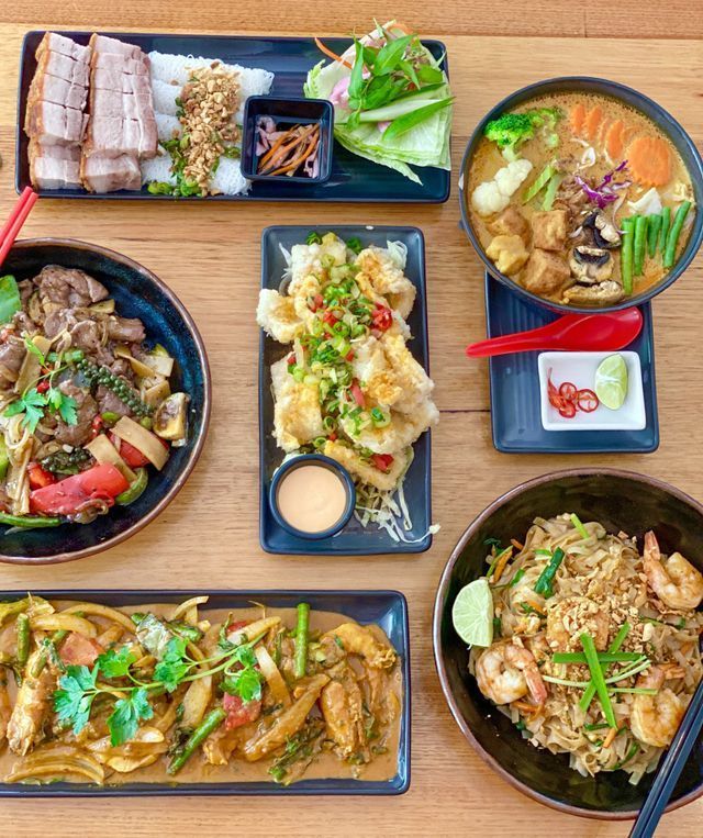 Overhead shot of various Southeast Asian dishes on a wooden table, including noodles, meats, and vegetables.