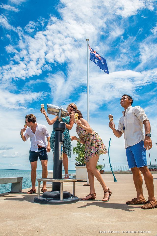 Four people eating ice cream while looking through a telescope next to an Australian flag on a sunny day.
