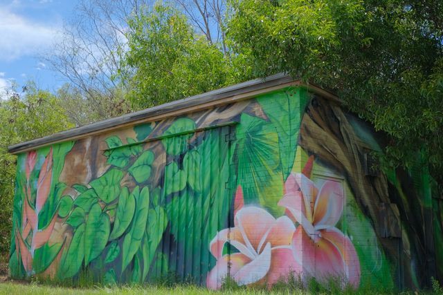 Mural on a weathered metal shed: green foliage and pink flowers against a cloudy sky.