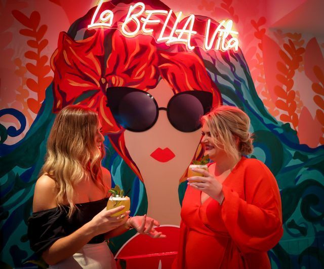 Two women with cocktails in front of a colorful wall mural. Neon sign reads 