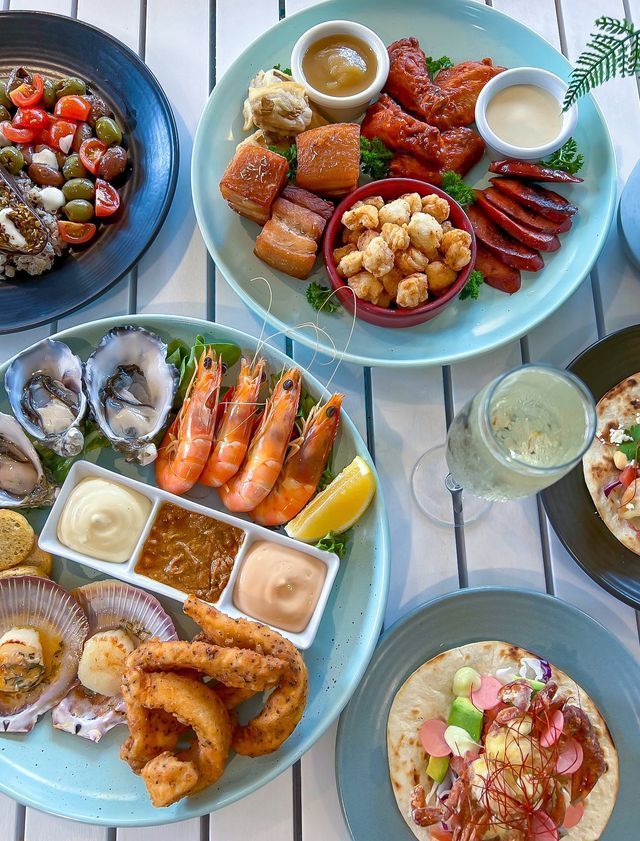 Platters of seafood, meats, and sides on a white table. Includes oysters, shrimp, and tacos.