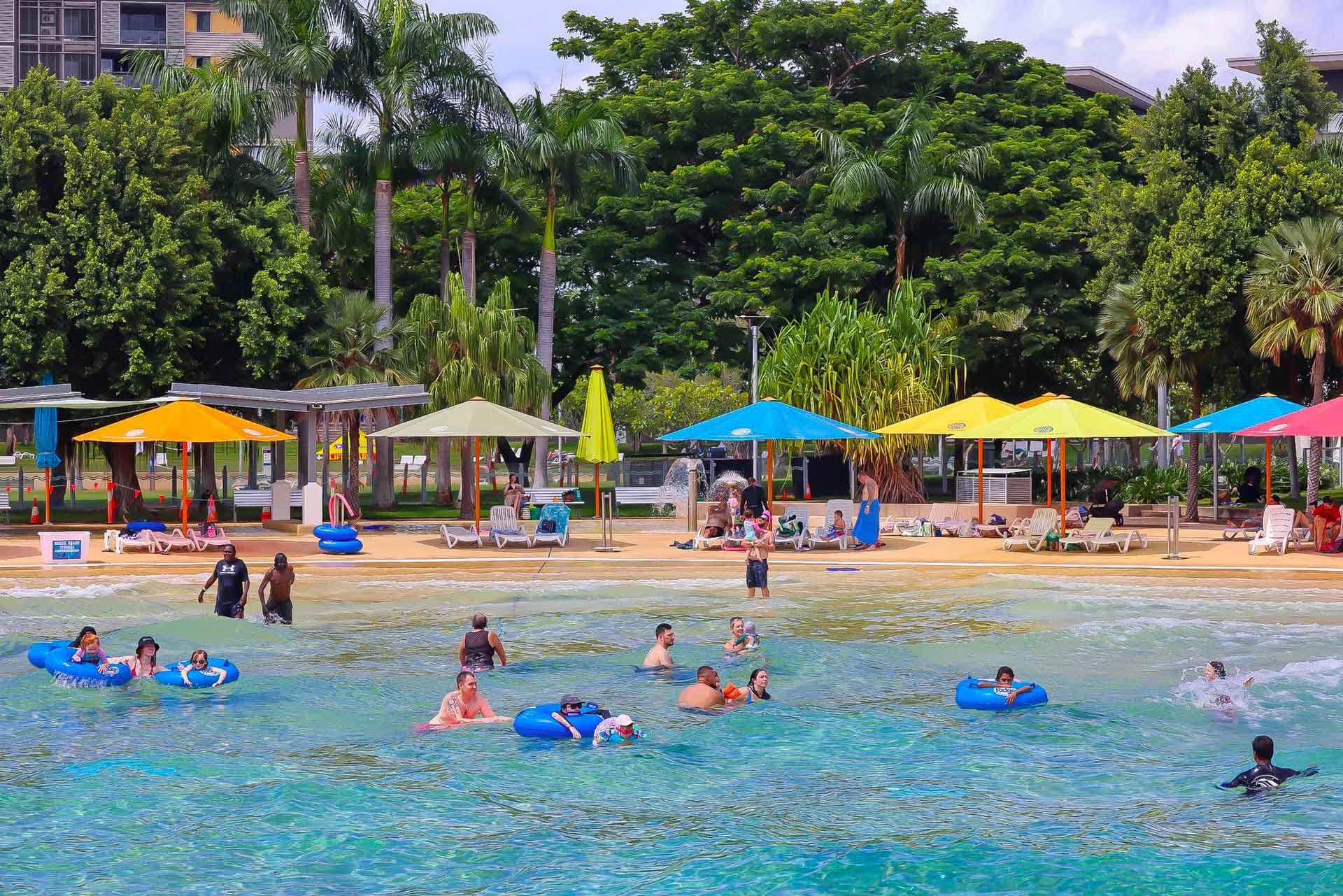 Beach scene with people swimming and relaxing under colorful umbrellas.