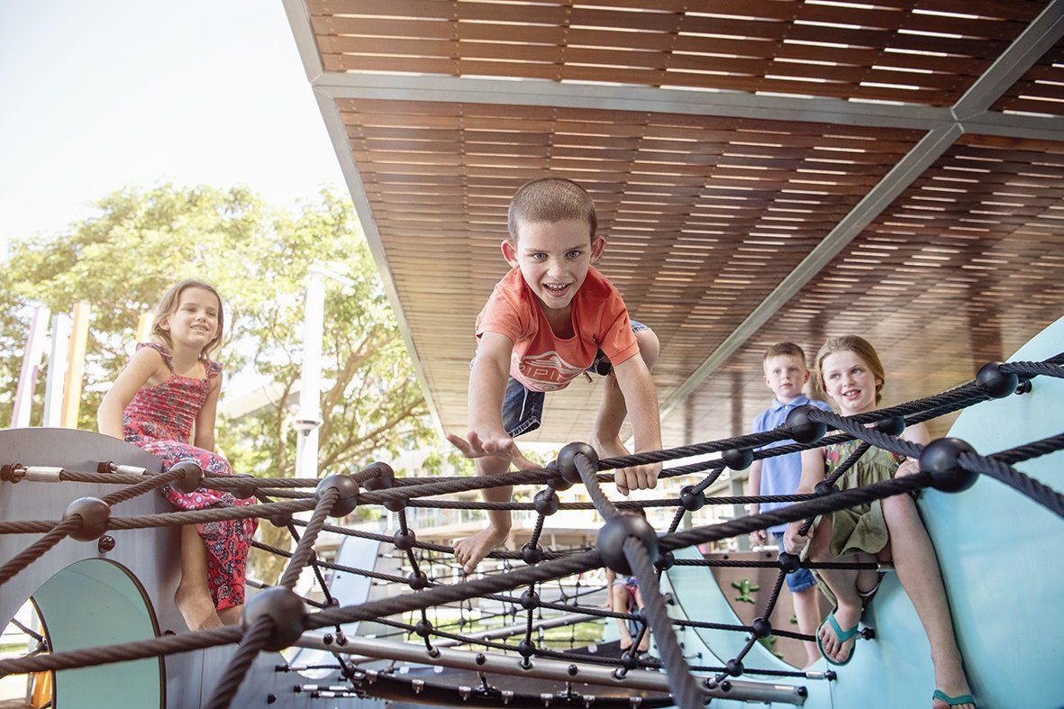 Children playing on a climbing structure at a park. One child is mid-climb, smiling. Sunny day.
