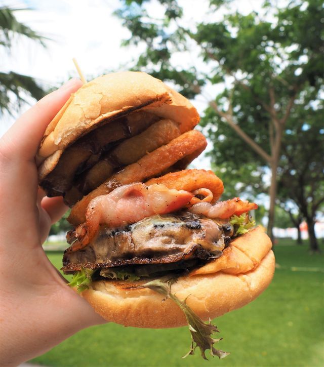 Person holding a large burger with onion rings, bacon, and mushrooms on a bun, outdoors.