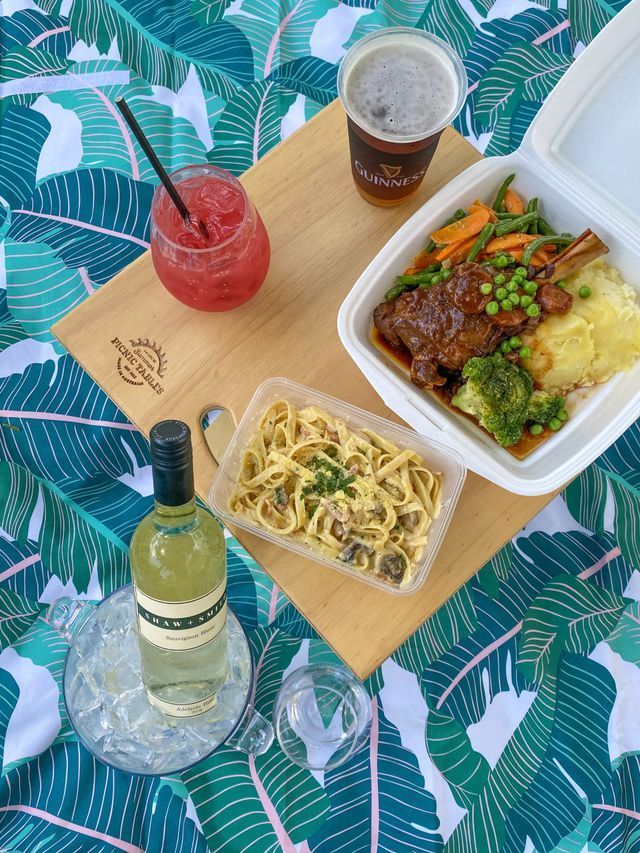 Picnic scene: lamb shank, pasta, mashed potatoes, vegetables, wine, beer, and a slushie on a blanket.
