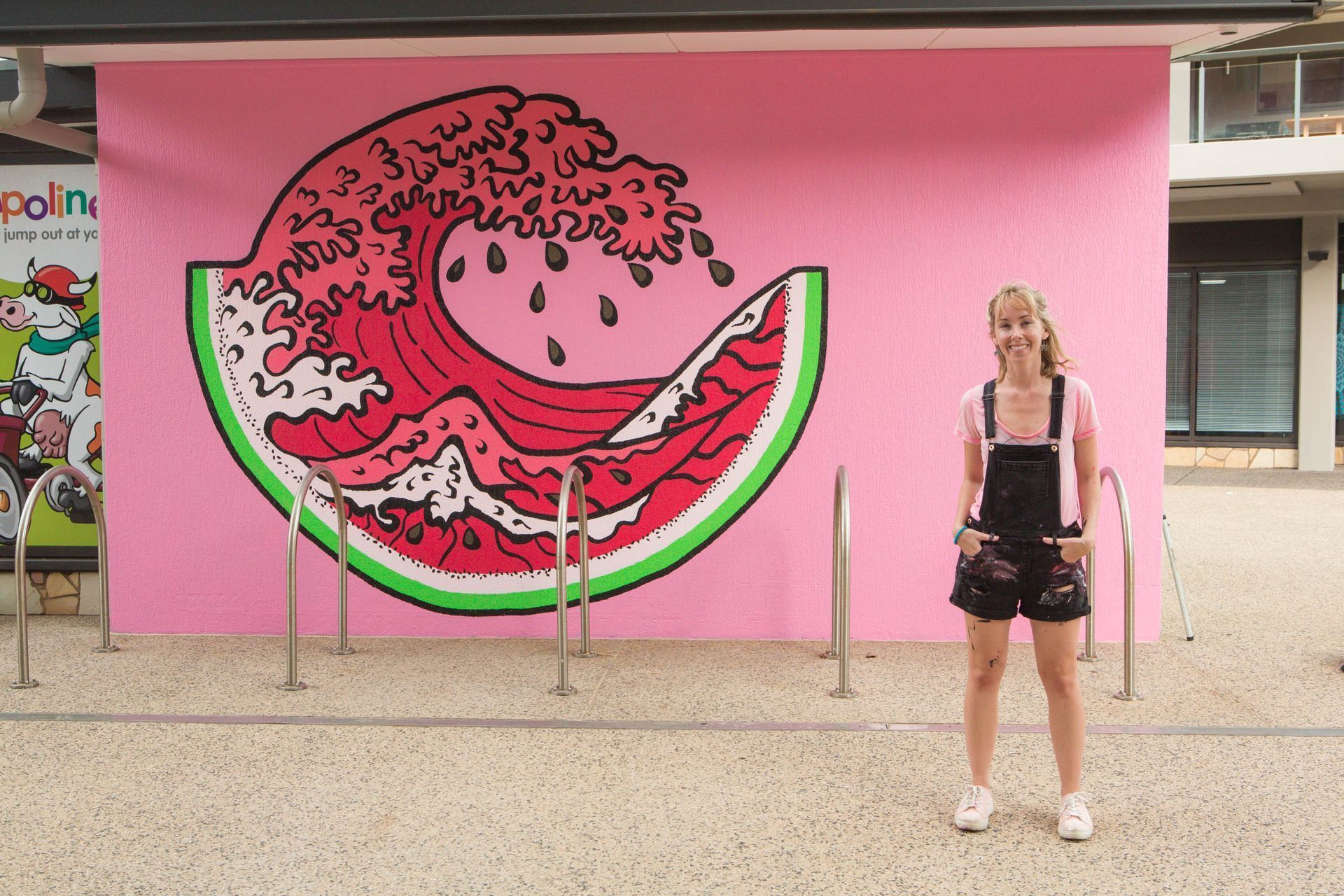 Woman standing in front of a watermelon wave mural. Pink background, black overalls.