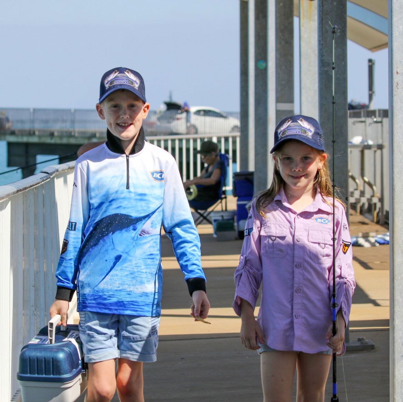 Two children wearing fishing attire walking on a pier, one carrying a tackle box and the other a fishing rod.