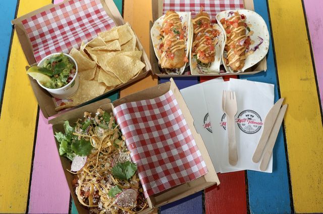 Mexican food spread: tacos, chips with guacamole, noodles, disposable cutlery, on a colorful table.