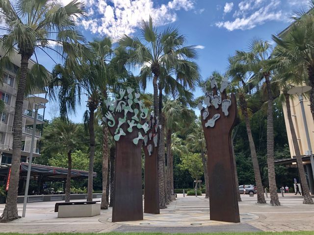 Tall metal sculptures in a plaza, framed by palm trees and buildings under a partly cloudy sky.