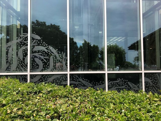 Glass windows with etched plant designs, reflecting trees and sky, above a green hedge.