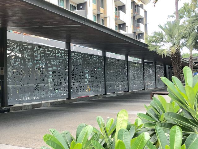 Long, patterned metal screen along a building's driveway, under a covered walkway, with greenery in the foreground.