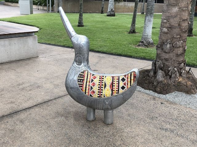 A metal bird sculpture with a colorful patterned back, outdoors on a concrete path.