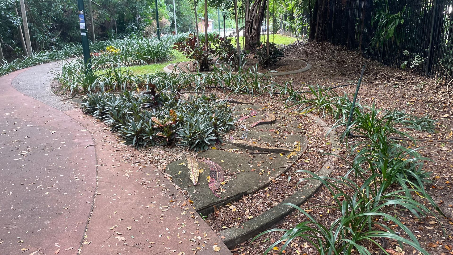 A curved paved path winds through a park with landscaping, including steps, plants, and fallen leaves.
