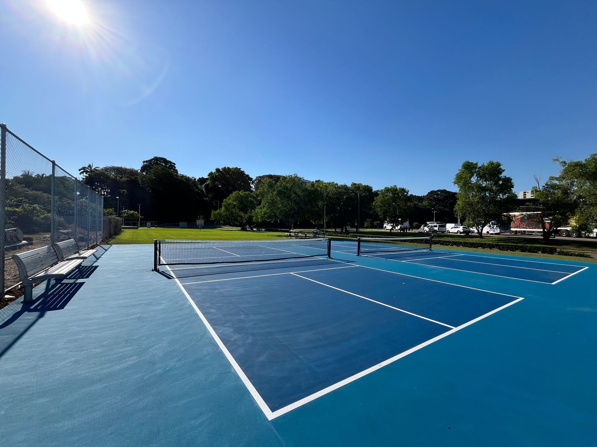 Outdoor pickleball courts on a sunny day. Blue courts with nets, surrounded by grass and trees.