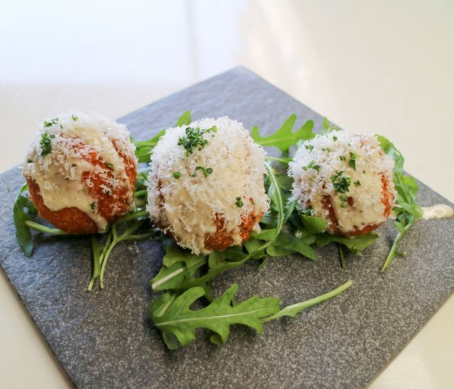 Three fried food balls topped with cheese and parsley, served on arugula, slate plate.