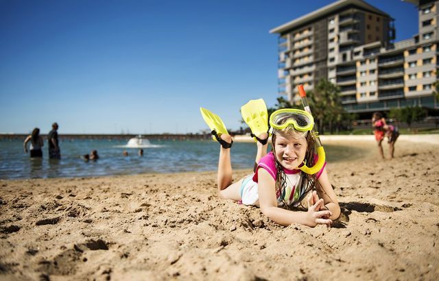 Girl in snorkel gear smiles on beach, legs in flippers, near water and buildings on a sunny day.