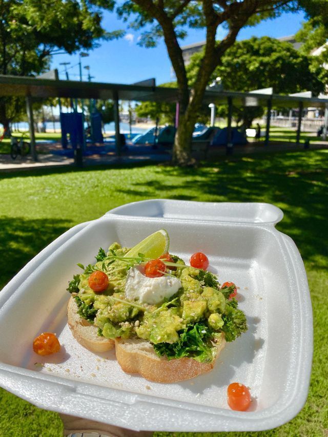 Avocado toast with cherry tomatoes and lime wedge in a styrofoam container, outdoors in a park.
