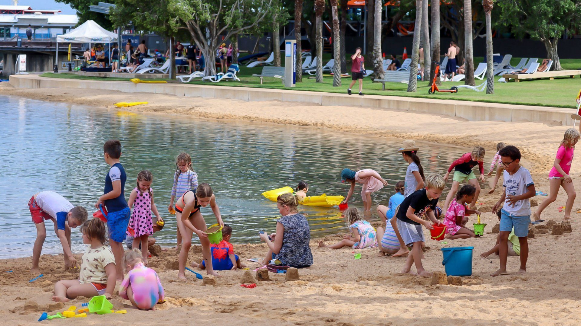 Children playing in sand at a beach-like area, some with buckets and shovels near water.