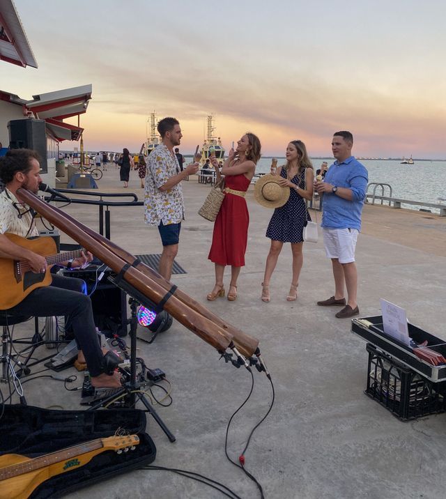 A musician playing didgeridoo and guitar for four people on a pier at sunset.