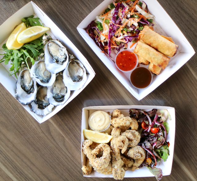Three takeaway boxes: oysters with lemon, spring rolls with salad, and fried calamari with sauce.