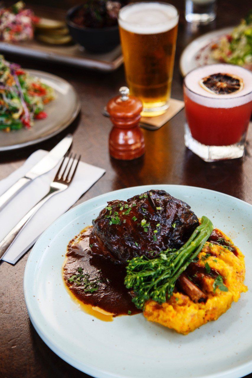 Plate with braised meat, mashed sweet potato, and greens, alongside drinks and other dishes.