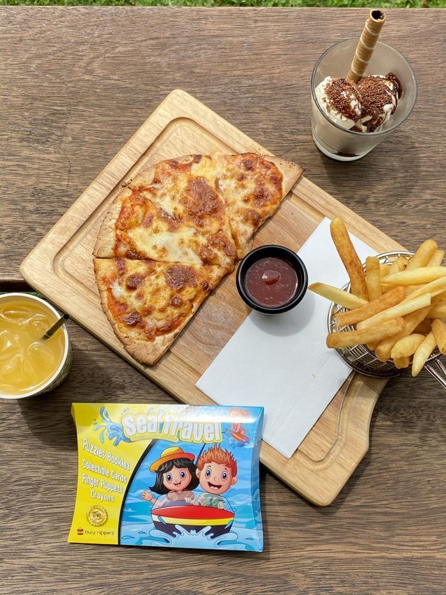 Pizza, fries, ice cream, juice, and a candy bag arranged on a wooden table, outside.