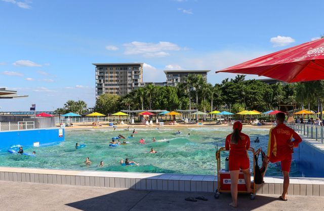 Wave pool with people swimming, lifeguards in red, and colorful umbrellas under a blue sky.