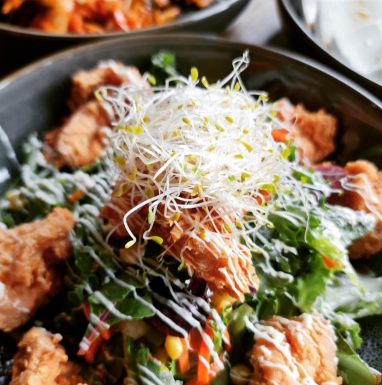 Salad bowl with fried chicken pieces, greens, vegetables, and sprouts, drizzled with white sauce.