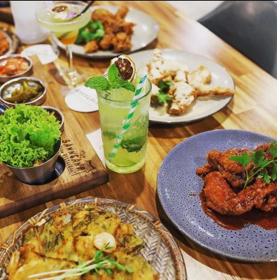 Table of food: Korean pancakes, fried chicken, wings, lettuce, and cocktails. Wooden table setting.