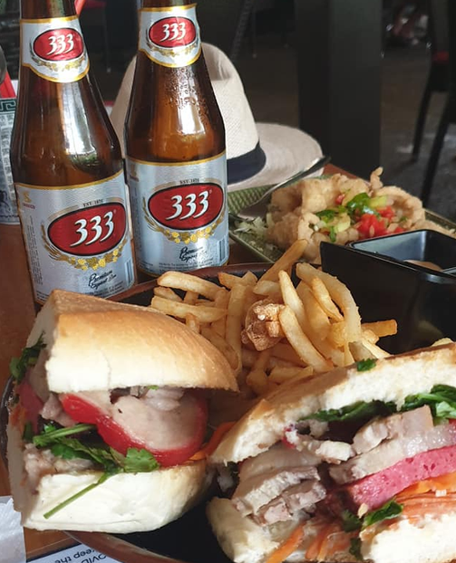 Two 333 beer bottles beside two banh mi sandwiches, fries, and fried food.