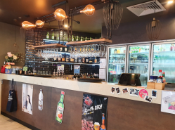 Bar interior with bottles, glasses, and decorative lights. Posters and a cooler are visible.