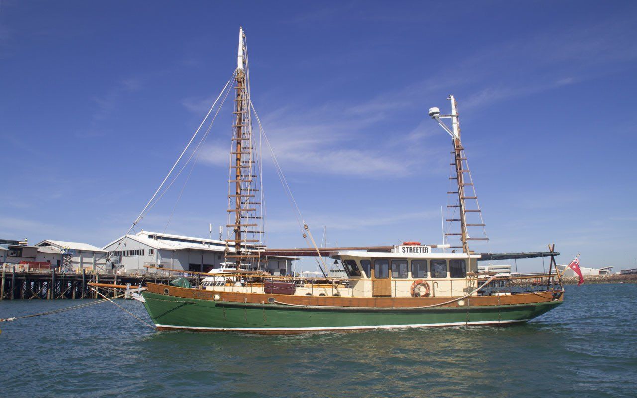 Green and brown boat with masts on the water near a pier under a blue sky.
