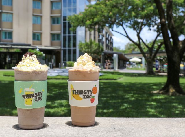 Two smoothies in branded cups on a ledge with a grassy background and buildings.