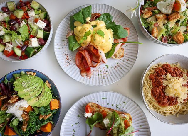 Overhead shot of various dishes: eggs benedict, pasta, salads on a white table.