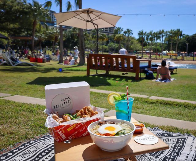 Picnic setup: fried chicken, noodles with egg, drink, on a grassy park by a pool.
