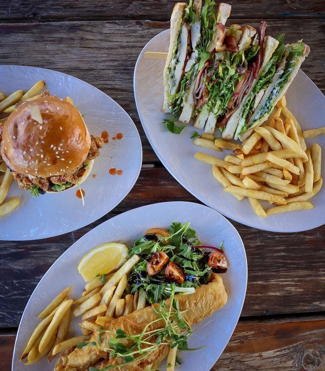 Plates of food: burger, club sandwich, and fish and chips with fries, salad, and lemon on a wooden table.