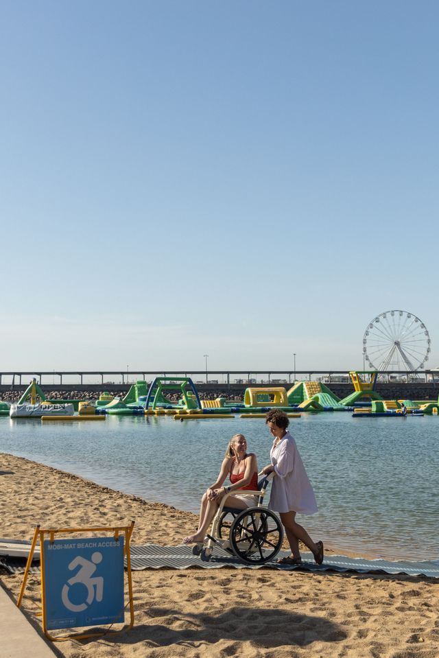 Woman in wheelchair at beach, being assisted by another. Accessible ramp, water park, ferris wheel in background.