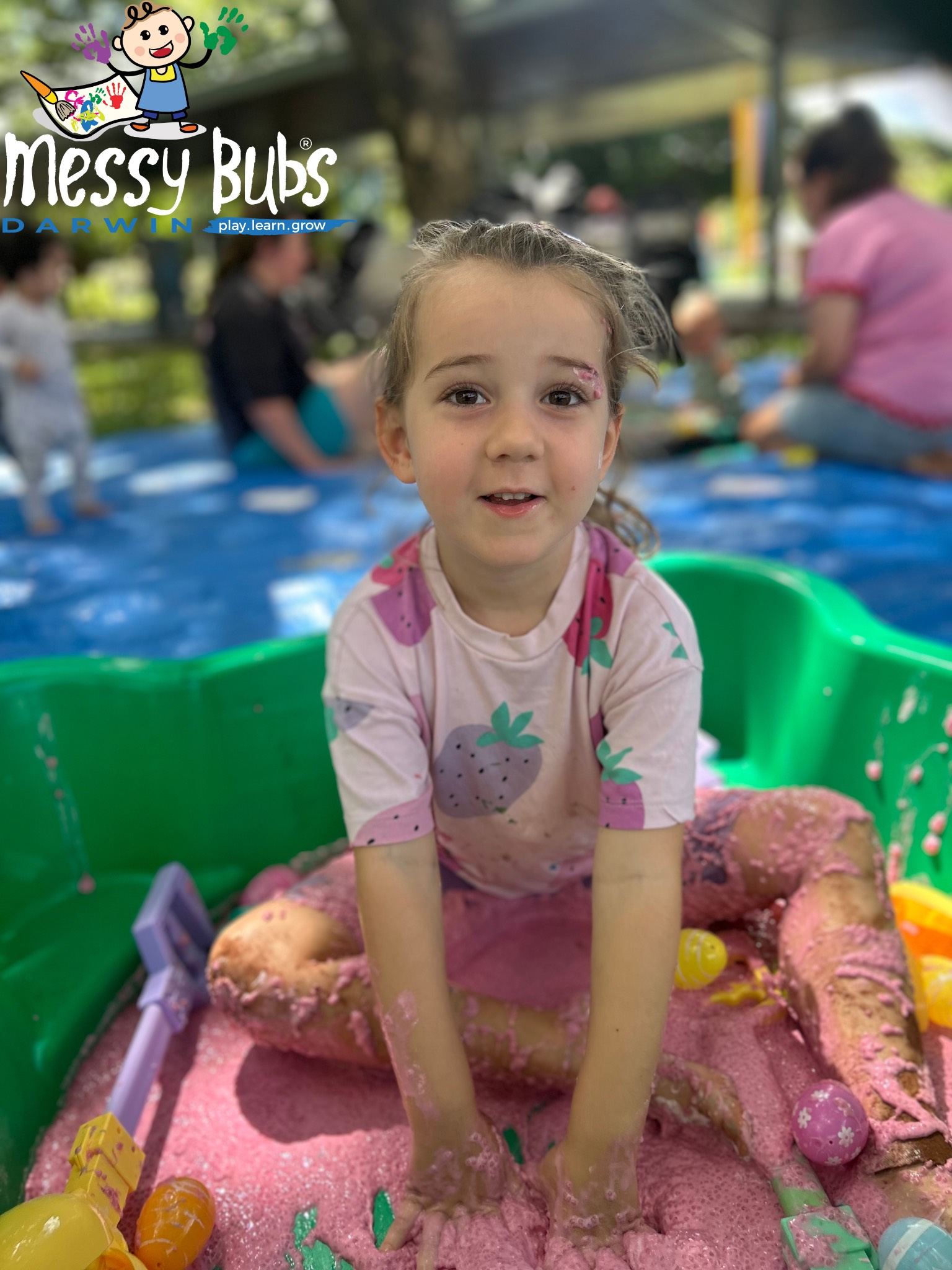 Girl in pink shirt sits in a green tub filled with pink, sparkly substance, smiling.