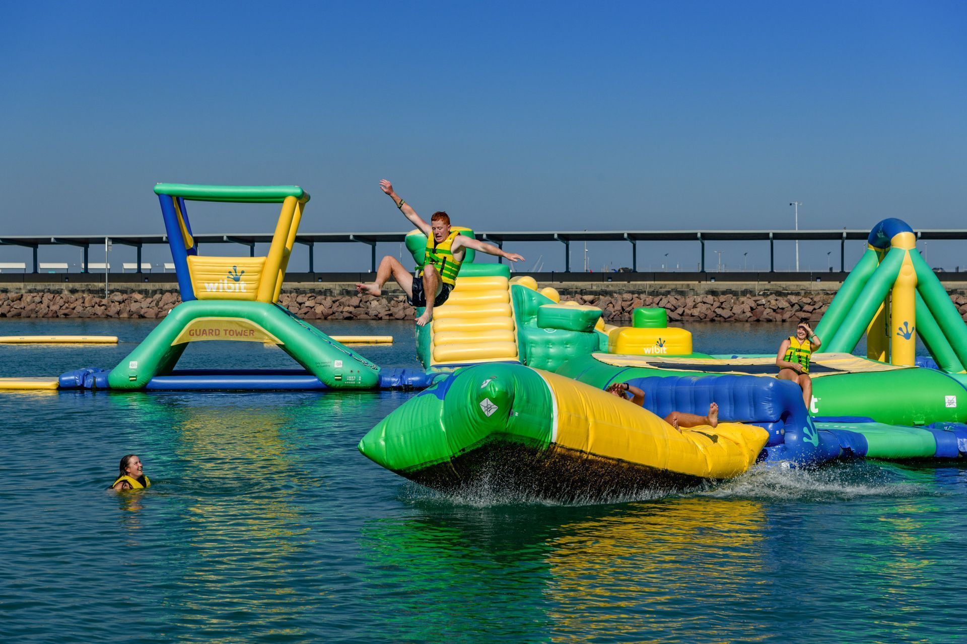 People playing on an inflatable water park. Brightly colored obstacles in blue water, sunny day.