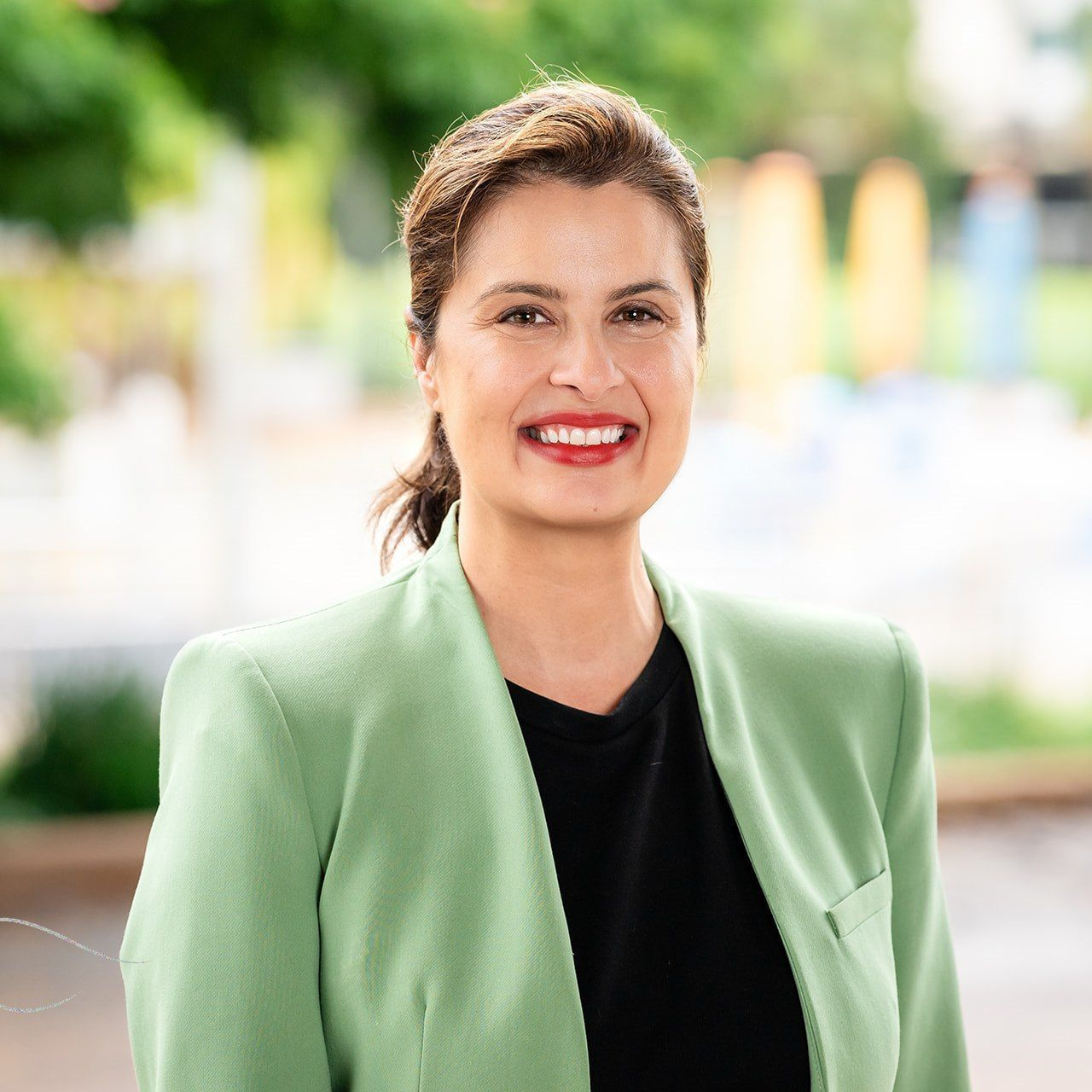 Woman with red lipstick wearing a green blazer, smiles, outdoors.