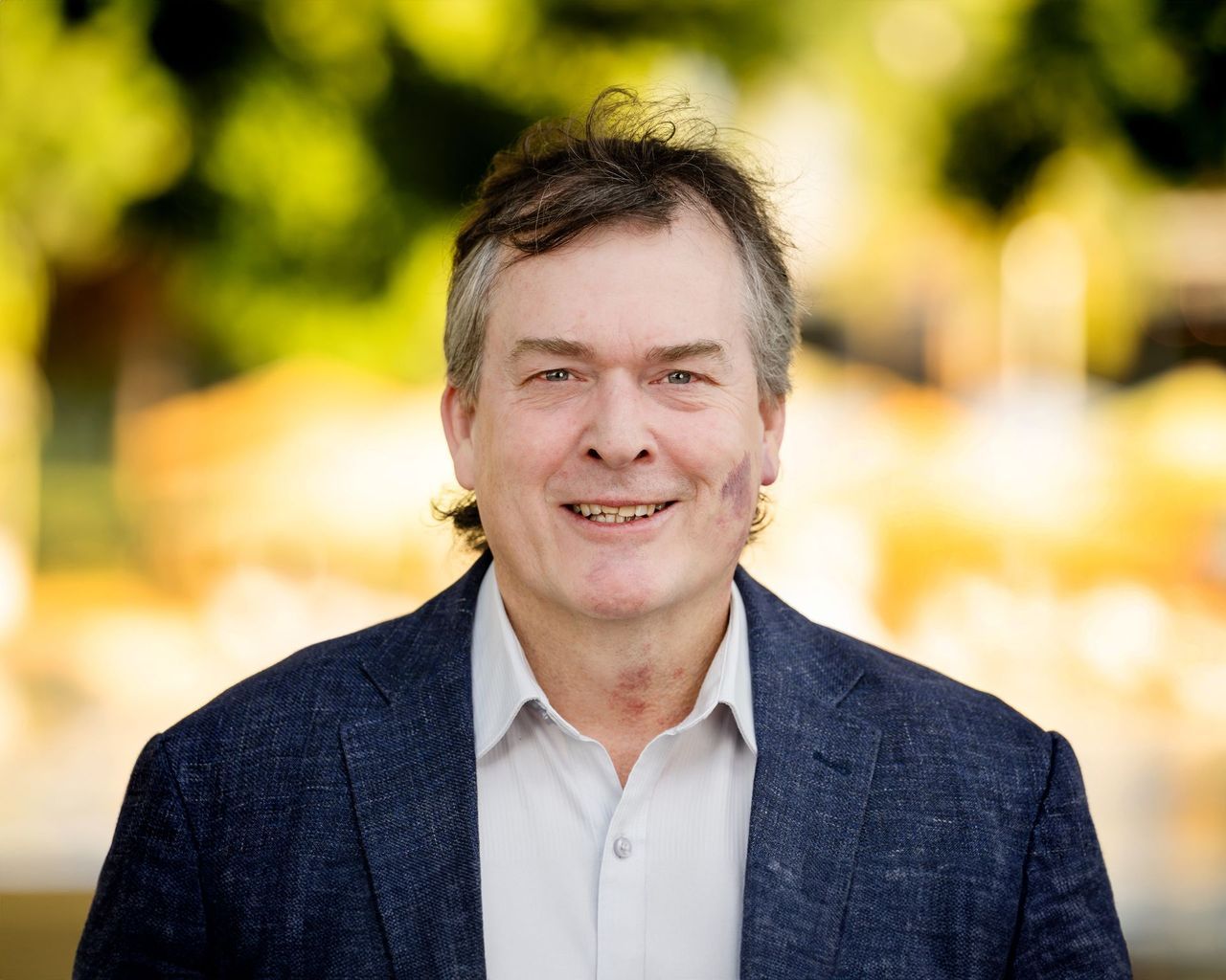Man in a blue blazer smiles outdoors; blurred green and yellow background.