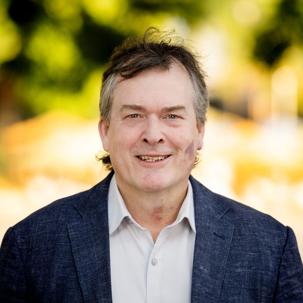 Man with brown hair smiles, wearing a navy blazer and white shirt, outdoors with blurred greenery.