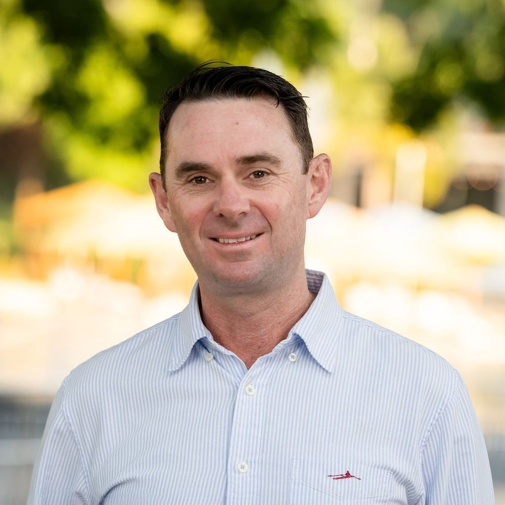 Man in light blue striped shirt smiles outdoors with blurred background.