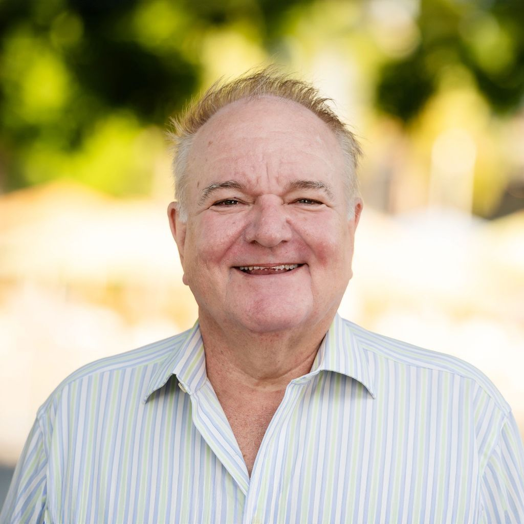 Smiling man with light skin, wearing a striped shirt, outdoors with blurred greenery.