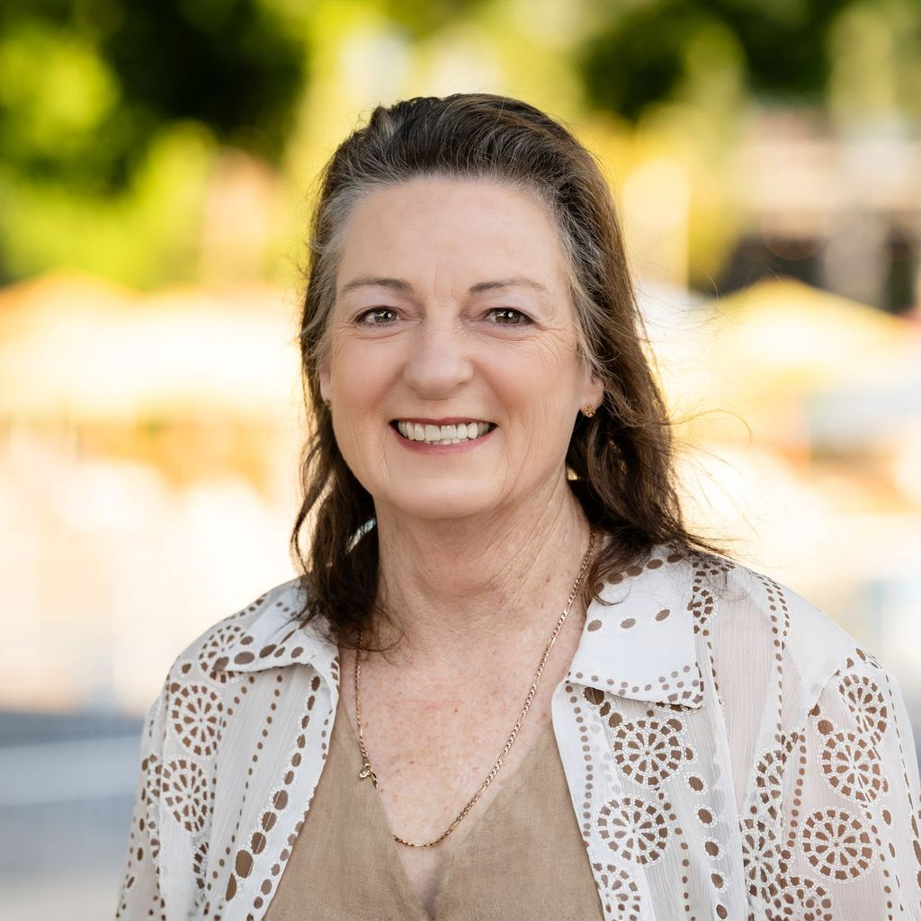 Woman with brown hair smiles, wearing a cream, open-front jacket. Outside with blurred background.