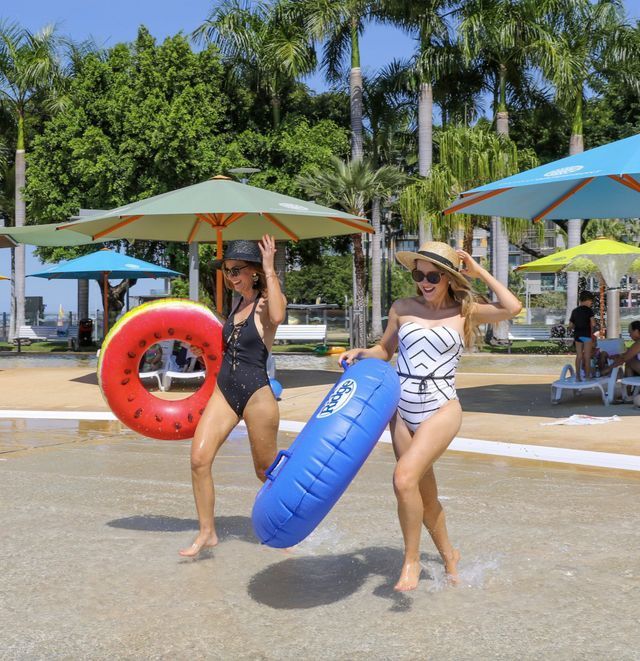 Two women in swimsuits at a water park, carrying inflatable tubes, smiling. Green trees and colorful umbrellas in background.